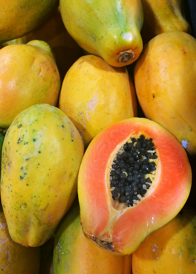 small, round halved Sunset Solo Papaya resting on top of several whole papayas - skin is deep yellow with specks of green - salmon colored flesh surrounds oval cluster of shiny black seeds