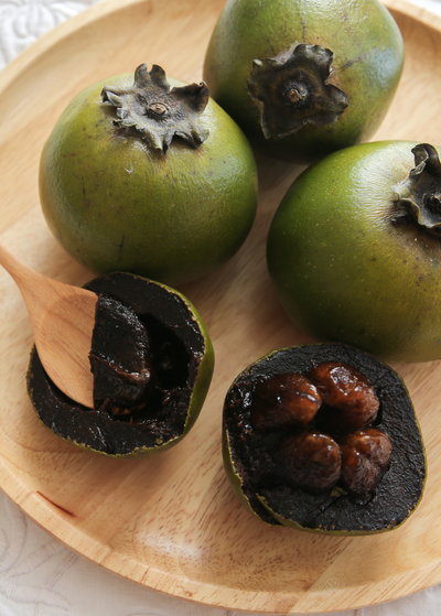 four Reineke Black Sapote brownish green  fruits on wooden plate - one is cut in half exposing black pudding like textured flesh and four seeds that look similar to dates - wooden spoon is scooping some of the fruit out